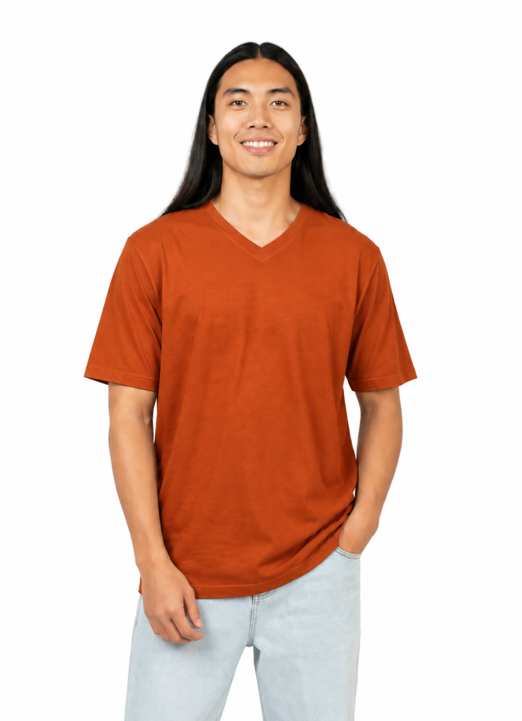 A man with long black hair wearing a rust-orange v-neck t-shirt and light blue jeans, standing against a solid white background.