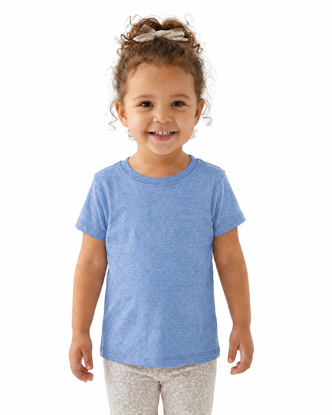 A young child with curly brown hair tied up with a bow is wearing a heathered light blue short-sleeve crewneck t-shirt and light beige patterned leggings, standing against a solid white background.
