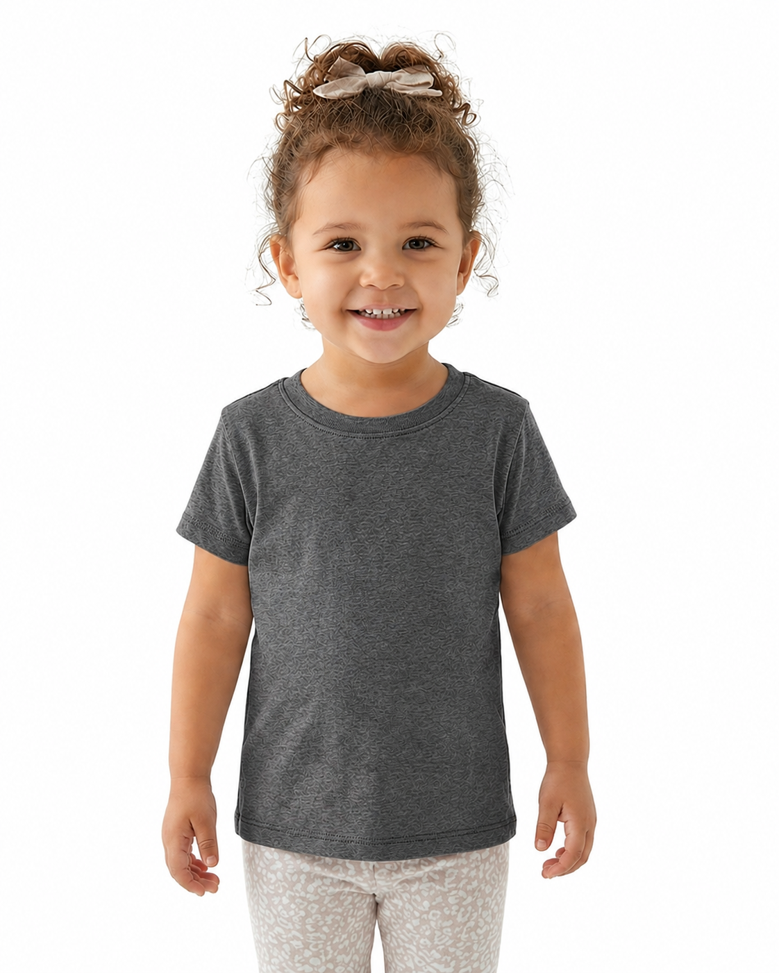 A young child with curly brown hair tied up with a bow, wearing a dark heather gray short-sleeve crewneck t-shirt and light patterned leggings, standing against a solid white background.