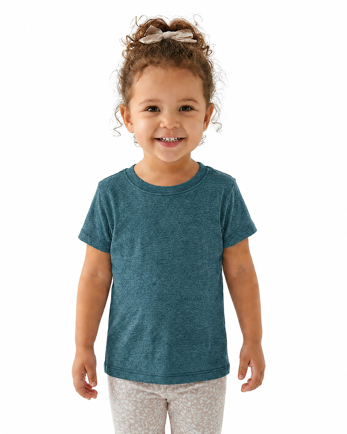 A young child with curly brown hair wearing a heathered teal blue short-sleeve t-shirt and light patterned pants, standing against a solid white background.