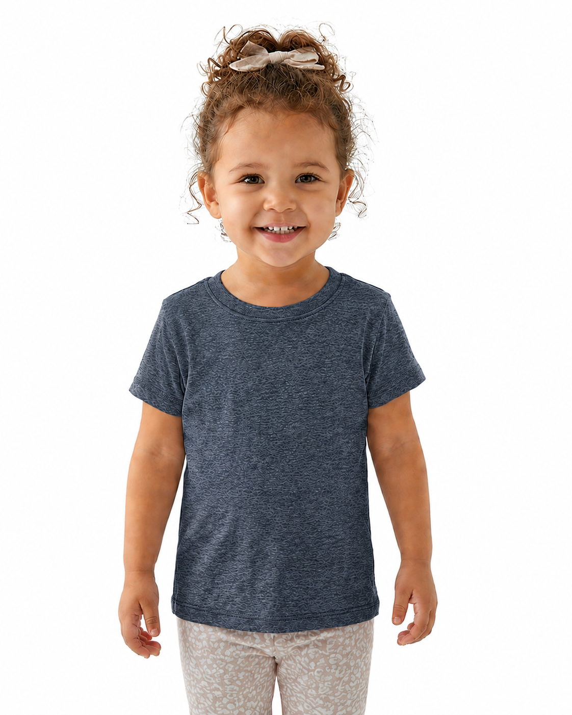A young child with curly brown hair wearing a heathered navy blue short-sleeve t-shirt and light patterned pants, standing against a solid white background.