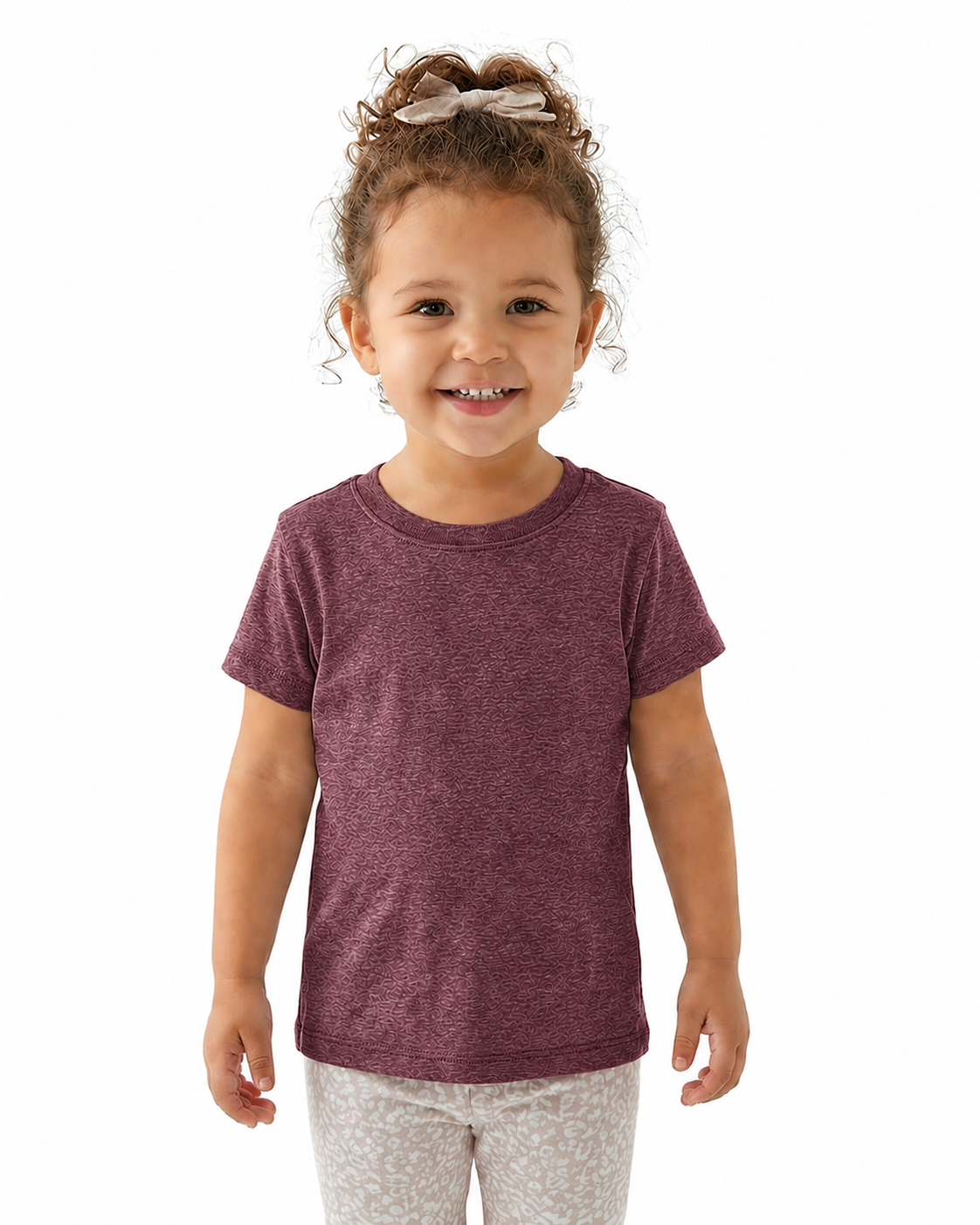 A young child with curly brown hair wearing a heathered burgundy short-sleeve t-shirt and light patterned leggings, standing against a solid white background.
