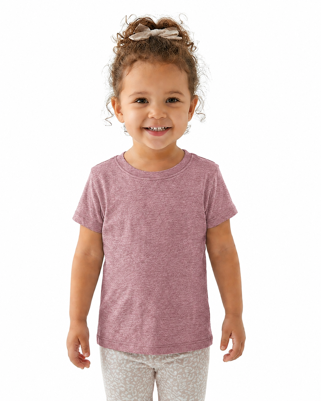 A young child with curly brown hair tied up with a bow, wearing a heathered dusty rose short-sleeve t-shirt and light patterned leggings, standing against a solid white background.