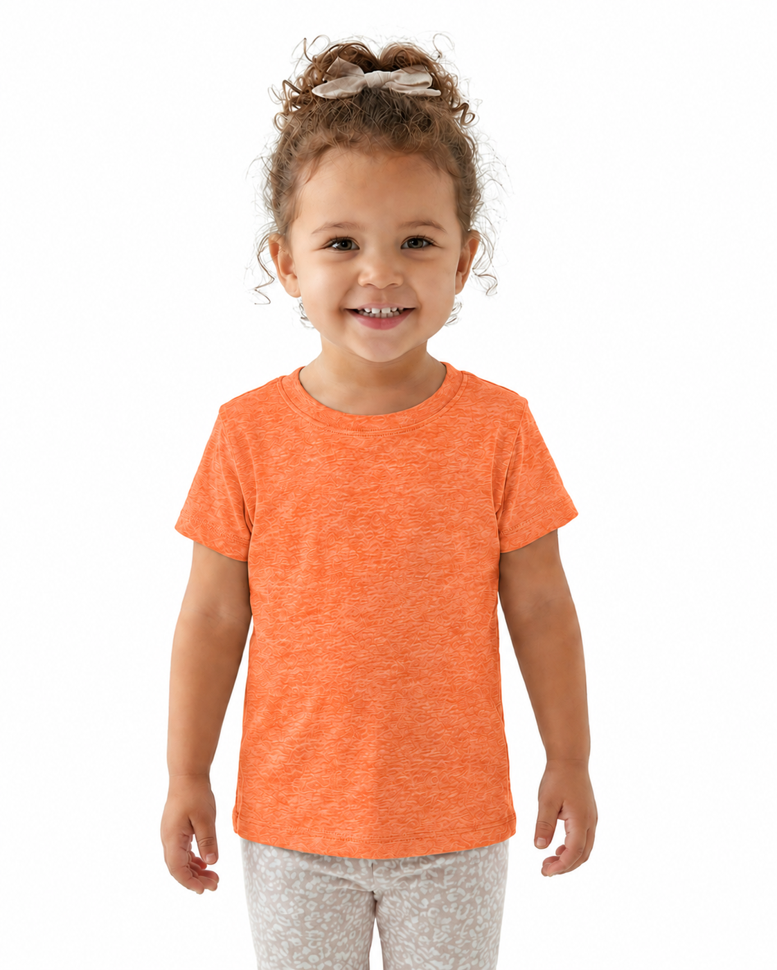 A young child with curly brown hair in a high ponytail wearing a heathered orange short-sleeve t-shirt and light gray patterned pants, standing against a solid white background.