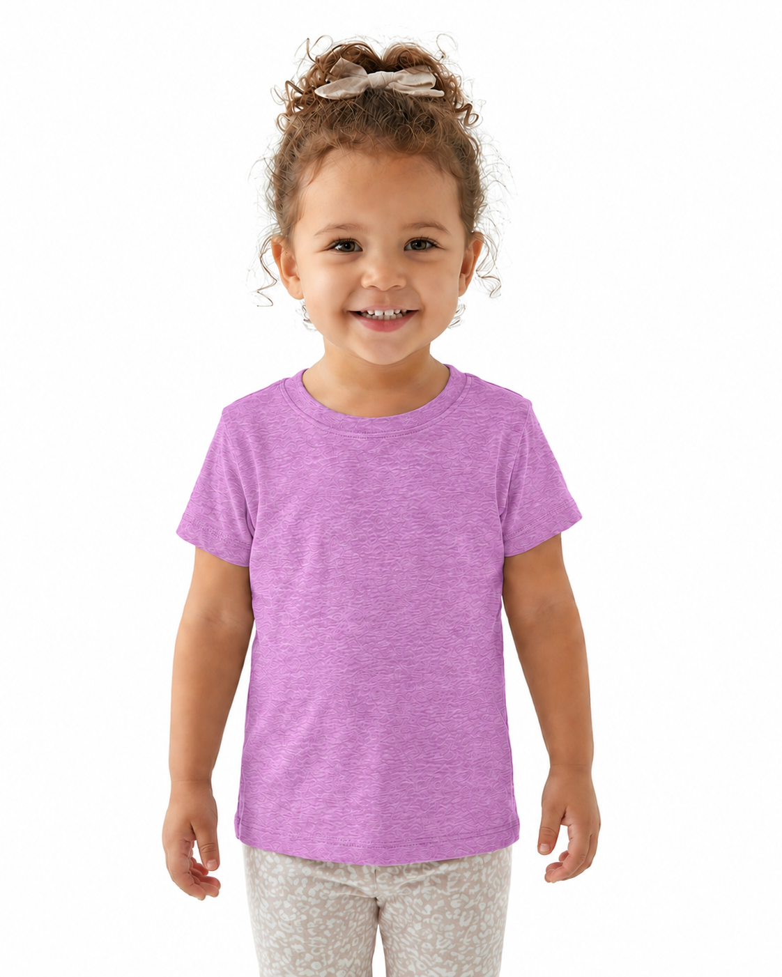 A young girl with curly brown hair in a high bun wearing a heathered light purple short-sleeve t-shirt and light patterned pants, standing against a solid white background.
