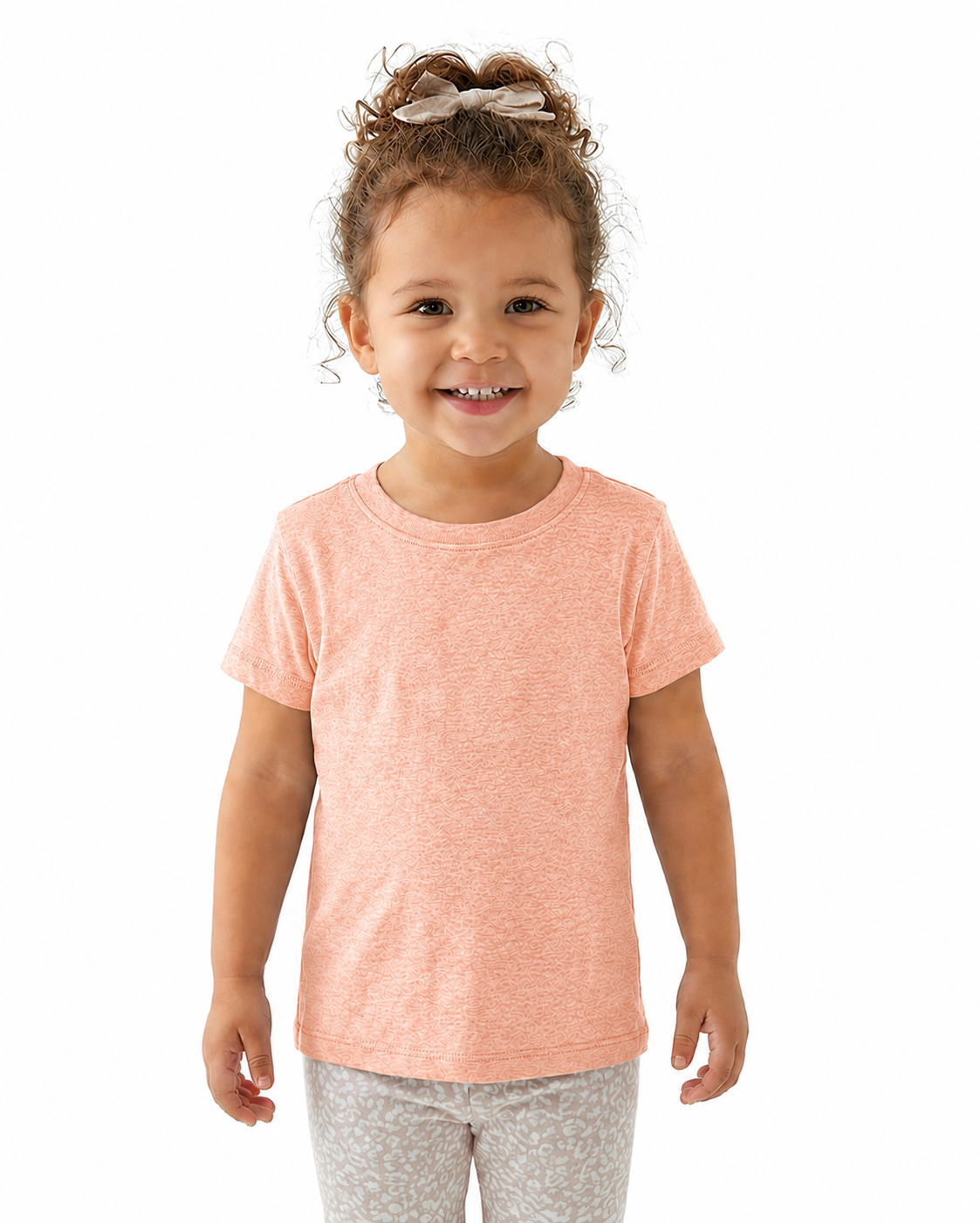 A young child with curly brown hair tied up with a bow, wearing a heathered peach short-sleeve t-shirt and light gray patterned leggings, standing against a solid white background.