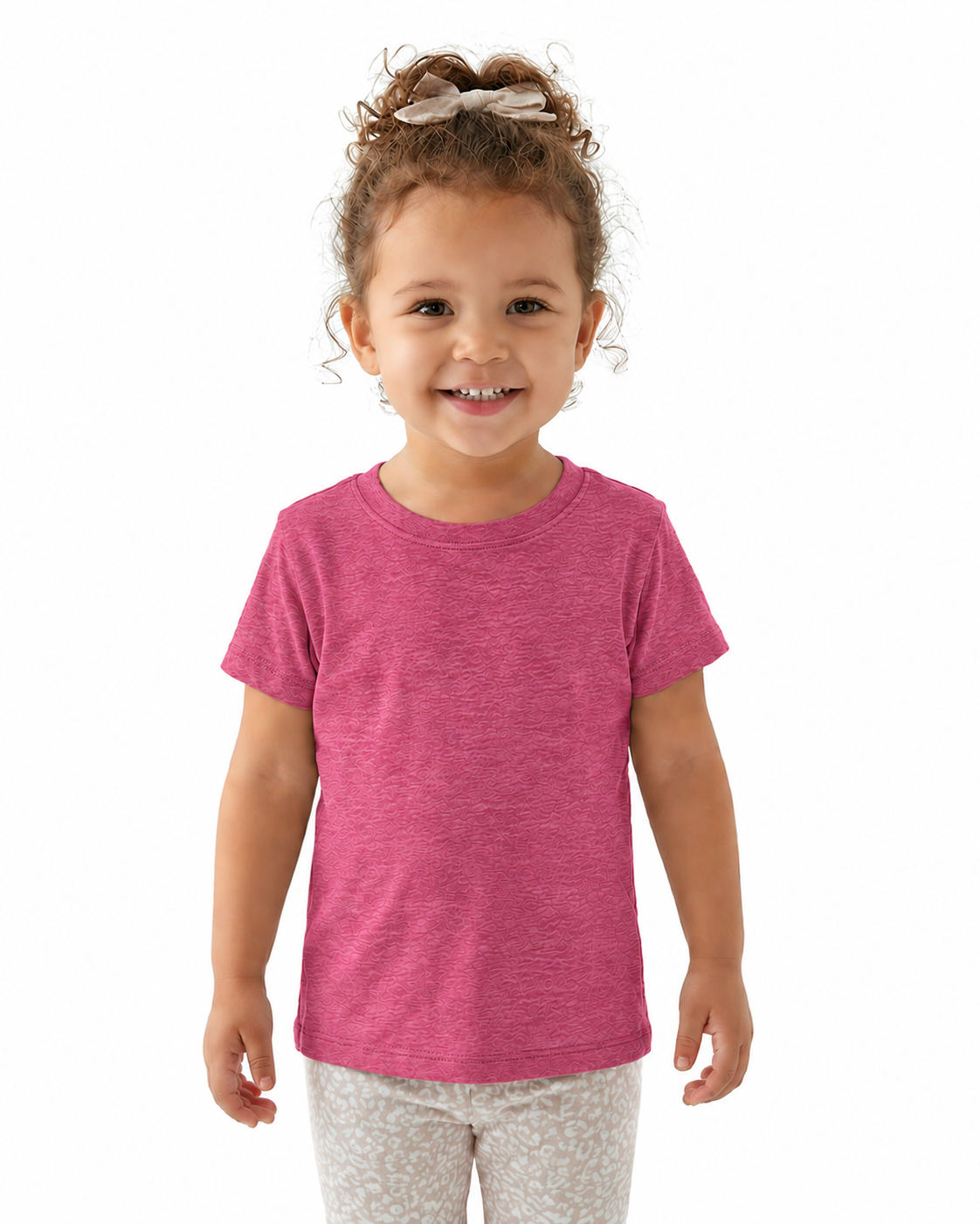 A young girl with curly brown hair tied up with a bow is wearing a heather pink short-sleeve t-shirt and light patterned leggings, standing against a solid white background.