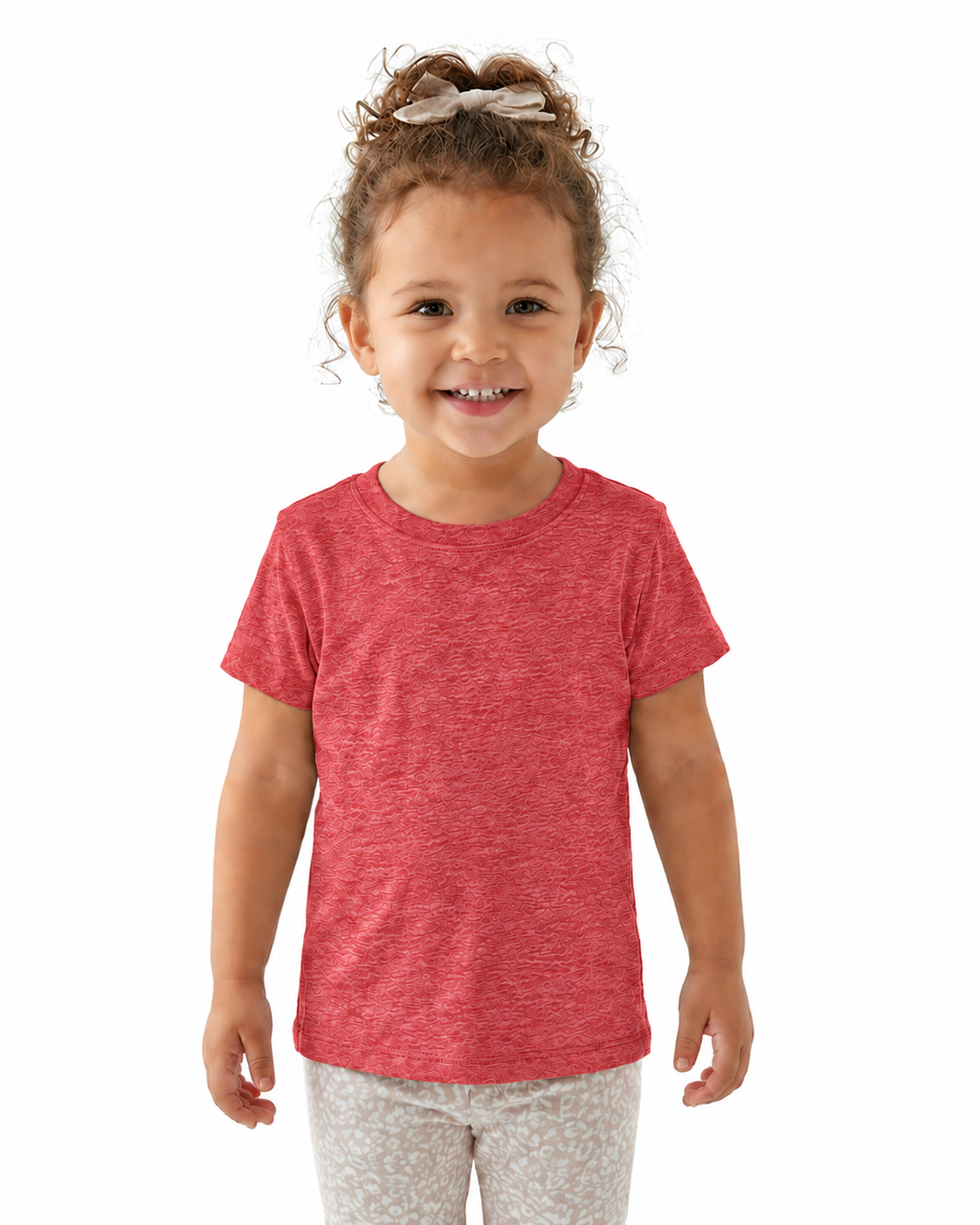 A young child with curly brown hair tied up with a bow, wearing a heathered coral-red short-sleeve t-shirt and light gray patterned pants, standing against a solid white background.