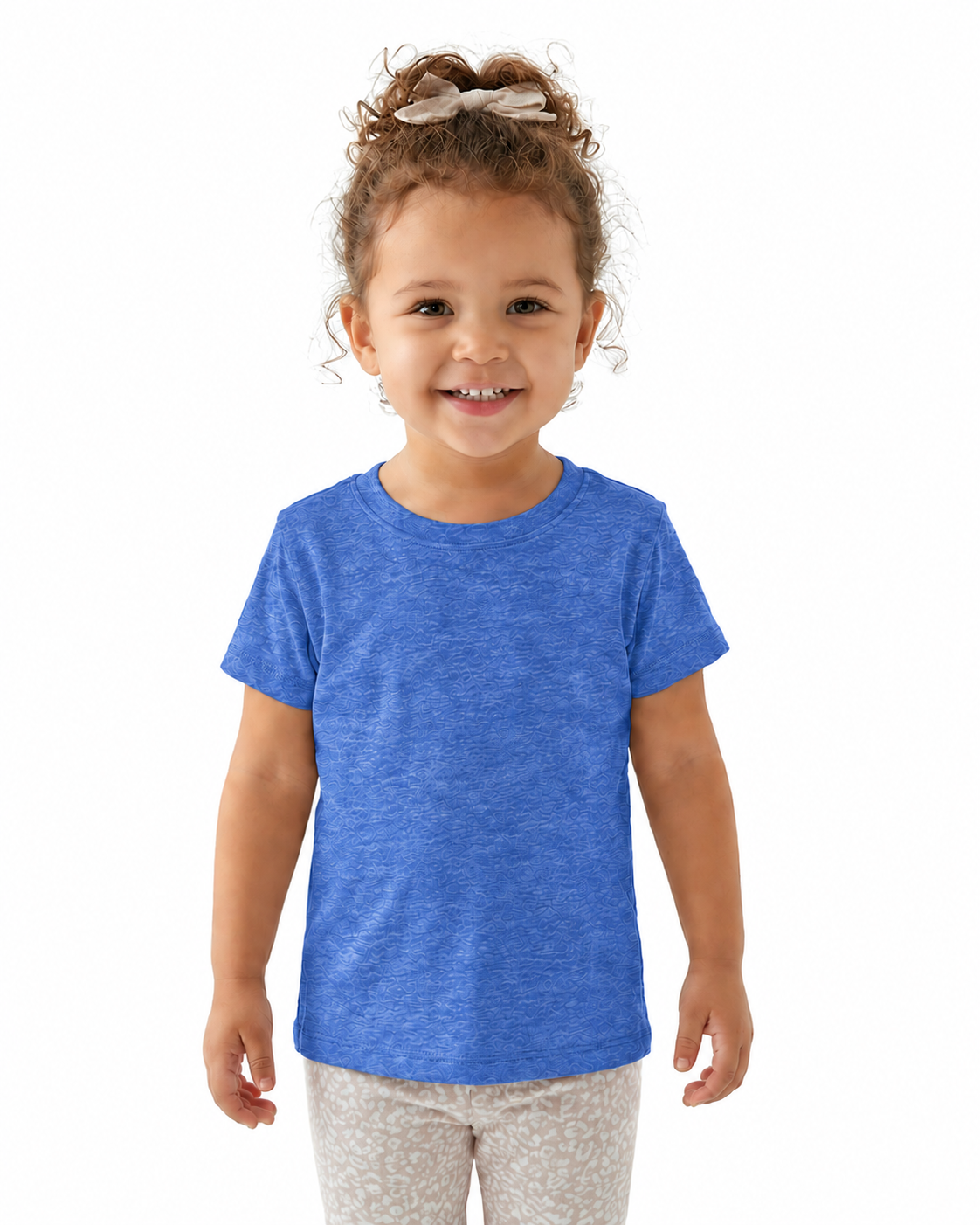A young child with curly brown hair tied up with a bow, wearing a heather blue short-sleeve crewneck t-shirt and light patterned pants, standing against a solid white background.
