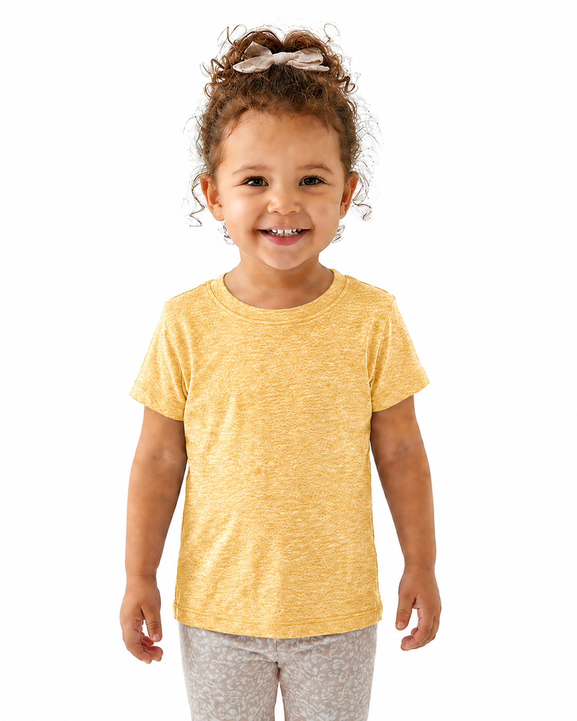 A young child with curly brown hair wearing a yellow heathered short-sleeve t-shirt and light gray patterned pants, standing against a solid white background.