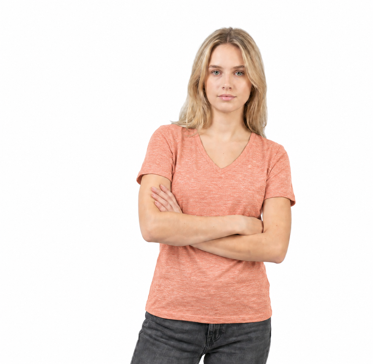 A woman with blonde hair wearing a heathered coral pink v-neck t-shirt and dark gray jeans, standing with her arms crossed against a solid white background.